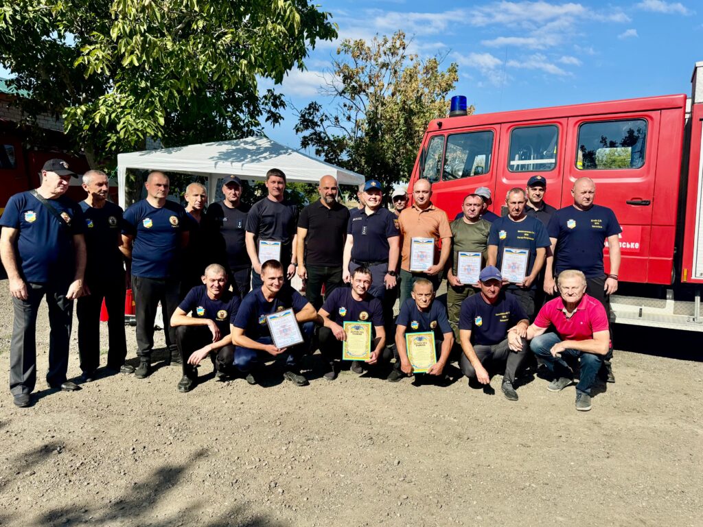 Близько двадцяти людей позують перед червоною пожежною машиною. Дехто тримає сертифікати або грамоти. Більшість — у формі, ймовірно рятувальники або пожежники. Позаду — дерева та намет. Атмосфера — урочиста, схожа на церемонію нагородження.