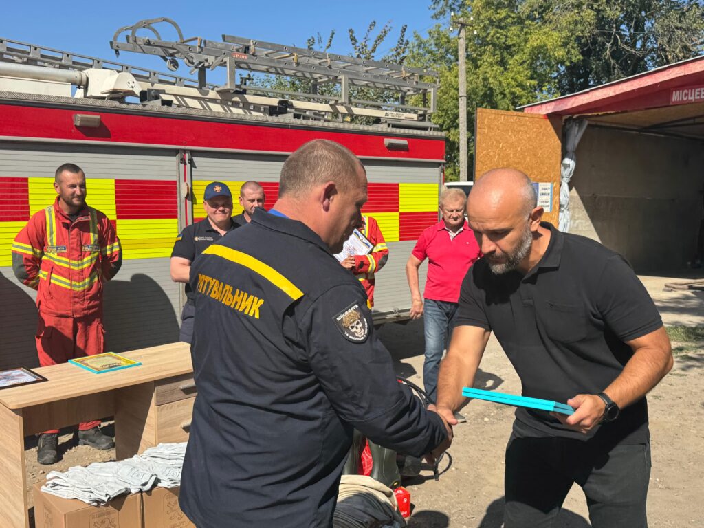 Група людей зібралася біля пожежної машини з червоно-жовтими смугами. Юліан Лучко вручає Подяку рятувальнику у формі з написом «РЯТУВАЛЬНИК» на спині. Поруч — люди в червоній формі, столи з документами та речами. Атмосфера — урочиста, пов’язана з нагородженням або передачею спорядження.