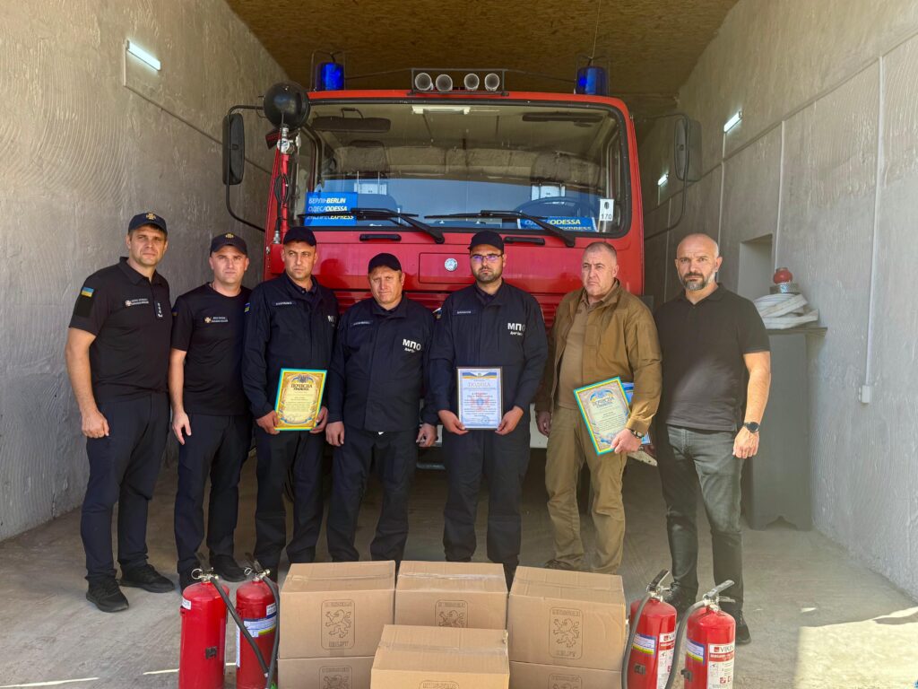 Сім дорослих стоять у гаражі перед червоною пожежною машиною. Четверо — у темній формі, один — у коричневій, двоє — у цивільному одязі. Троє тримають сертифікати. На підлозі — картонні коробки та чотири червоні вогнегасники. Атмосфера — офіційна, схожа на вручення нагород або передачу обладнання.