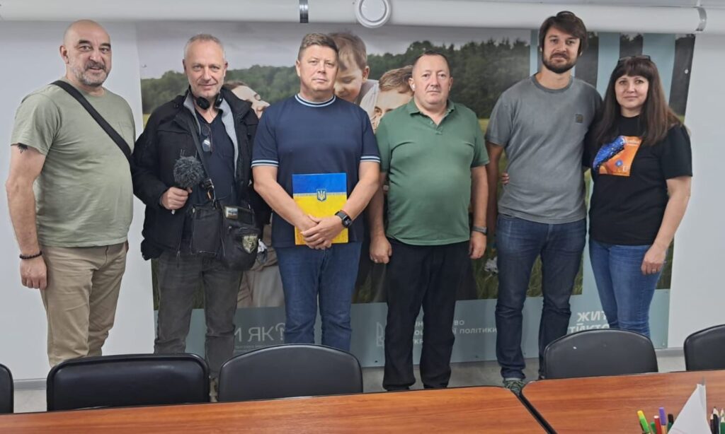 🏛️ Альтернативний текст (стислий): Група людей бере участь у нараді в кімнаті з синім банером «Херсонська обласна військова адміністрація». За столом — учасники в формі, цивільному одязі та традиційному українському вбранні. На столі — документи, пляшки з водою, іменні таблички. Центральна постать веде обговорення.