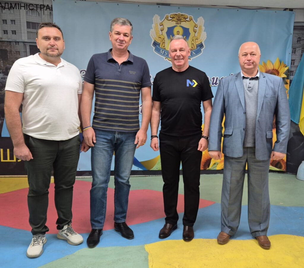 Чотири дорослі особи стоять перед банером із гербом України, соняшниками та синьо-жовтим прапором. Люди одягнені у повсякденний та напівофіційний одяг. Під ногами — яскраві геометричні фігури.