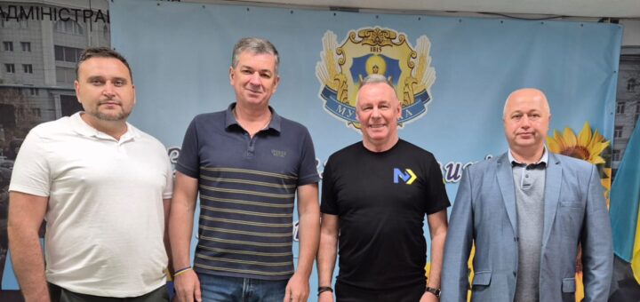 Чотири дорослі особи стоять перед банером із гербом України, соняшниками та синьо-жовтим прапором. Люди одягнені у повсякденний та напівофіційний одяг. Під ногами — яскраві геометричні фігури.