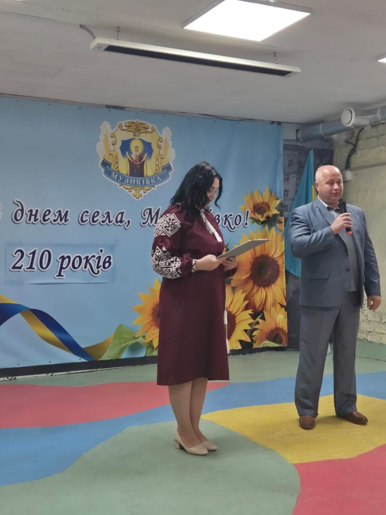 Михайло Жадан та ведуча стоять на яскраво розфарбованій сцені під час святкування. Позаду — банер із соняшниками, написом «З днем села, Мушкатівка! 210 років» та стрічкою в кольорах українського прапора. Обоє тримають мікрофони.