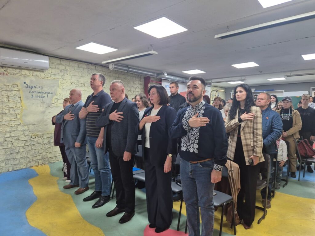 Група людей стоїть у приміщенні з правими руками на серці — момент патріотизму або вшанування. На стіні напис: «Україна починається з тебе!». Видно ряди стільців і яскраво розфарбовану підлогу.