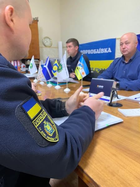 Троє людей сидять за дерев’яним столом у службовому кабінеті. Один — у формі ДСНС України, з нашивкою «Україна» на рукаві. На столі — мініпрапорці, документи, записники та календар із написом «LEAD». Позаду — банер із назвою «ЧОРНОБАЇВКА» та іншими українськими словами.