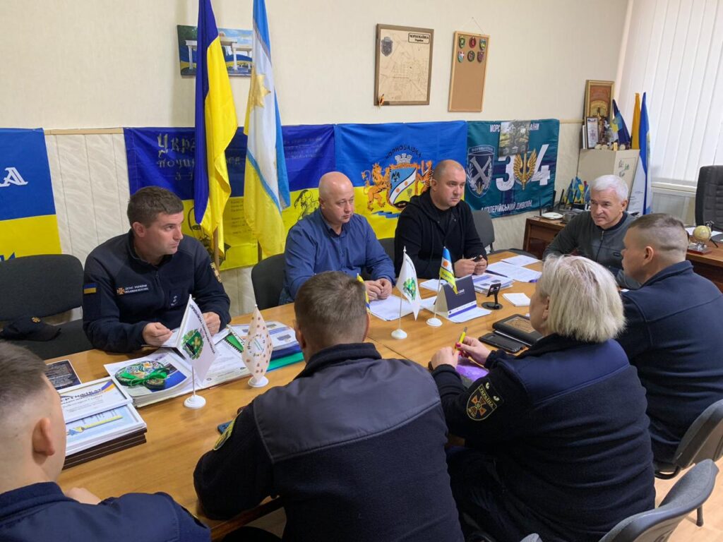 Вісім людей у темній формі сидять за прямокутним конференційним столом. На столі — документи, теки, іменні таблички та мініпрапорці (зокрема український). На стіні — прапори, емблеми, карти, сертифікати та портрет. Атмосфера — офіційна, пов’язана з надзвичайними ситуаціями або службовою координацією.