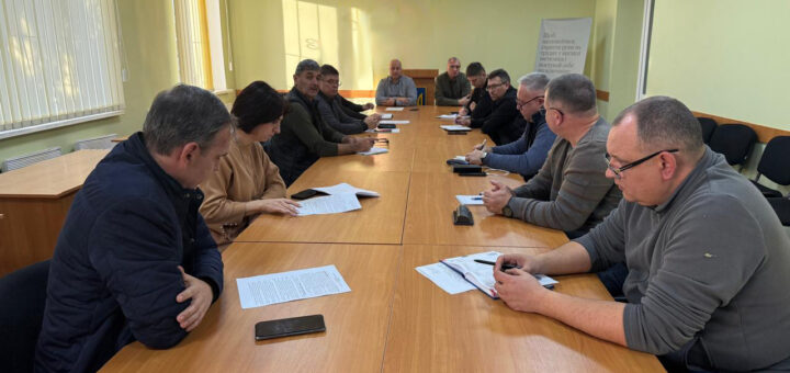 П’ятнадцять людей сидять за довгим столом у світлій кімнаті з українським прапором і гербом на стіні. На столі — документи, телефони, ручки.