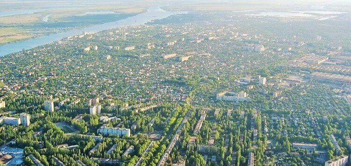 Аерофотозйомка міста з щільною забудовою. Видно житлові та комерційні будівлі, між якими розташовані зелені зони та вулиці, обсаджені деревами. У верхній частині зображення — велика річка, що плавно вигинається через ландшафт. Загальний вигляд демонструє поєднання міського планування з природним середовищем.