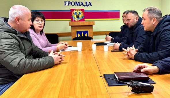 У залі засідань за дерев’яним столом сидять шестеро людей. На стіні позаду — напис українською «МУЗИКІВСЬКА СІЛЬСЬКА ТЕРИТОРІАЛЬНА ГРОМАДА», над яким розміщений герб України. Під написом — трибуна з кольоровим емблемою та синьо-жовтим оформленням. На столі — блокноти, папери та чашки.