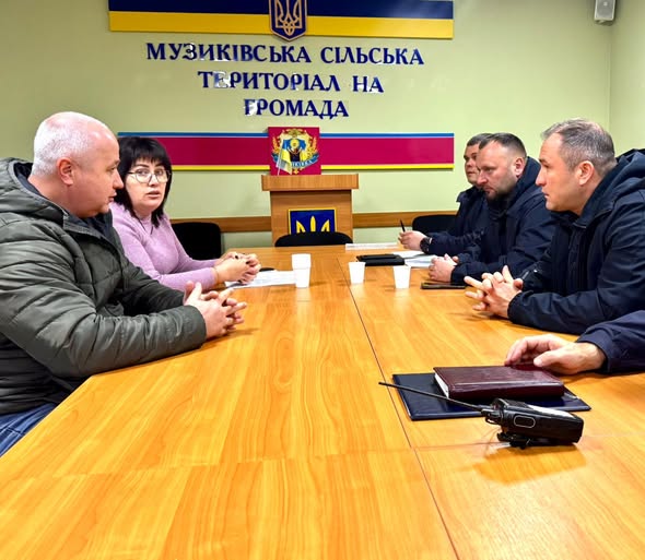 У залі засідань за дерев’яним столом сидять шестеро людей. На стіні позаду — напис українською «МУЗИКІВСЬКА СІЛЬСЬКА ТЕРИТОРІАЛЬНА ГРОМАДА», над яким розміщений герб України. Під написом — трибуна з кольоровим емблемою та синьо-жовтим оформленням. На столі — блокноти, папери та чашки.