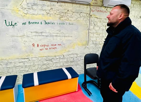 У яскравій кімнаті з розмальованою цегляною стіною стоїть чоловік, обличчям до стіни. На стіні — написані кольоровими літерами українські патріотичні фрази, зокрема «Ще не вмерла в Україні і слава і воля...» та «Я в серці маю те, що не вмира...». Поруч — лавки з синіми та білими подушками і чорний стілець. Підлога пофарбована в червоний, синій і жовтий — кольори українського прапора.