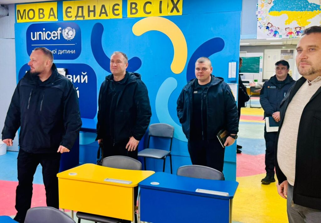 У яскраво оформленому класі стоїть група чоловіків. Приміщення має сині та жовті парти — кольори українського прапора. На стіні — абстрактні фігури та логотип ЮНІСЕФ. Над ними — банер із написом українською «МОВА ЄДНАЄ ВСІХ».