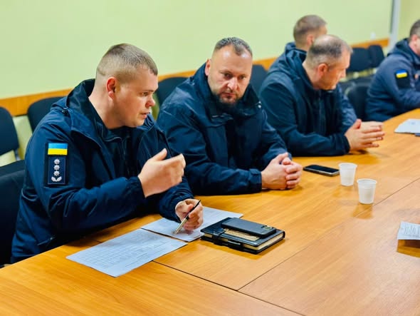 У кімнаті для нарад за дерев’яним столом сидить група людей у формі з нашивками українського прапора. Один із них, ліворуч, жестикулює ручкою, ніби пояснює щось. На столі — документи, смартфон, блокнот і одноразові стаканчики.