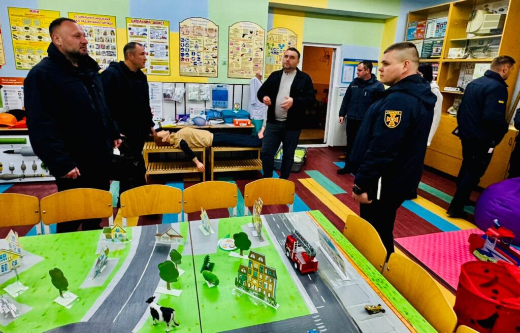 У навчальній кімнаті, ймовірно присвяченій цивільному захисту, перебувають кілька людей, частина з них — у формі з емблемами. На стінах — навчальні плакати українською мовою з темами «МІНИ», «ЦИВІЛЬНИЙ ЗАХИСТ» та іншими, що стосуються надзвичайних ситуацій. На лаві — медичні манекени та аптечки. На столі — детальна макетна модель містечка з дорогами, будівлями, деревами, машинами (зокрема пожежною) і коровою.