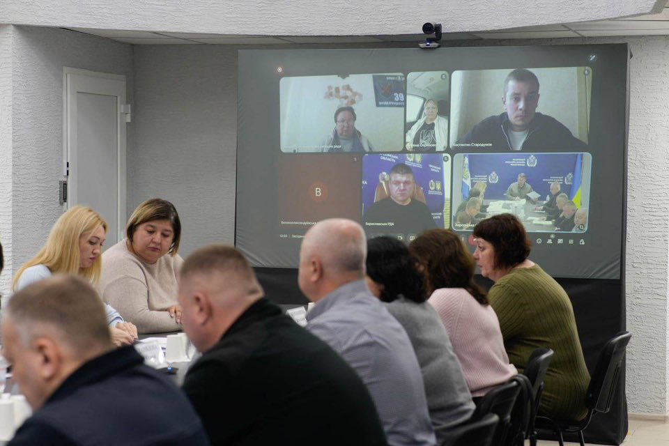 У конференц-залі люди сидять навколо столу, беруть участь у зустрічі. На задньому плані — великий екран, розділений на кілька вікон із учасниками відеоконференції. Деякі з них мають фони з прапорами або офіційною символікою.