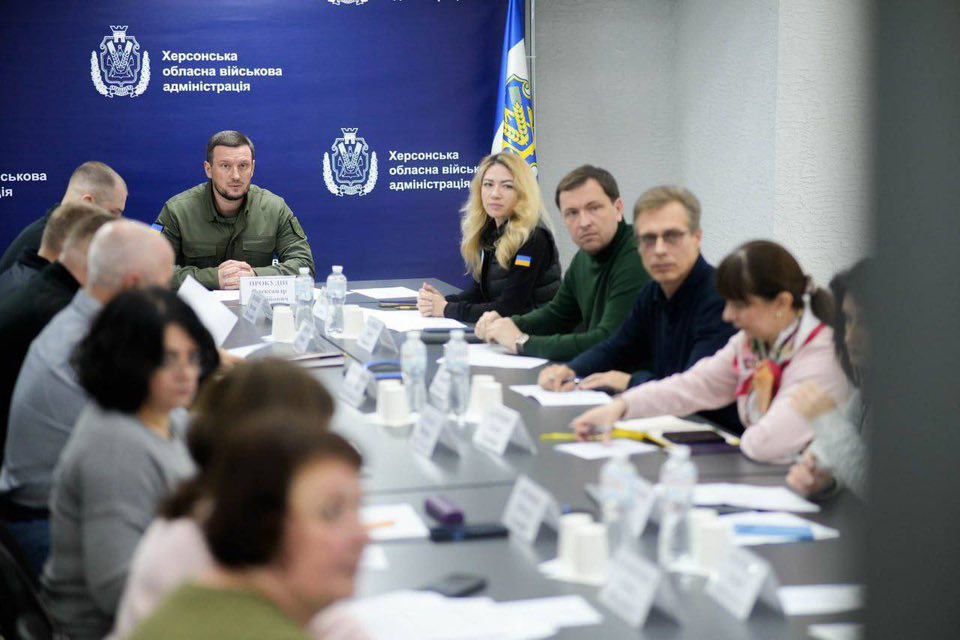 У кімнаті з довгим столом проходить офіційна зустріч. Учасники сидять із документами, табличками з іменами та пляшками води. На задньому плані — емблема та напис українською: «Херсонська обласна військова адміністрація». Видно прапор України.