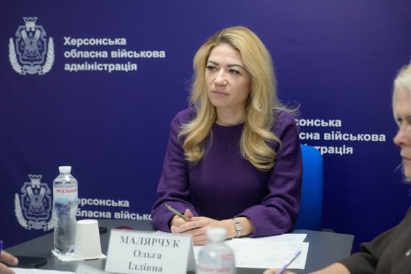 Жінка з довгим світлим волоссям сидить за конференційним столом. Вона вдягнена у фіолетовий верх. Позаду — синій фон із написом українською: «Херсонська обласна військова адміністрація». Перед нею — табличка з ім’ям: «МАЛЯРЧУК Ольга Іллівна». На столі — документи, ручка, пляшки з водою. 