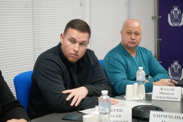 Двоє чоловіків сидять за конференційним столом під час офіційної зустрічі. На столі — пляшки з водою, чашки, документи та гучномовець. Позаду — фіолетовий банер з емблемою та кириличним текстом.