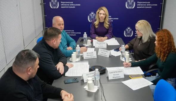 Шестеро людей сидять за конференційним столом у приміщенні з написом на фоні: «Херсонська обласна військова адміністрація». У кожного — іменна табличка, документи, пляшки з водою та мікрофони.