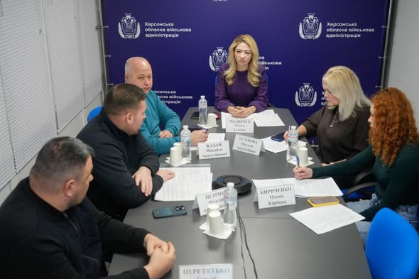 Шестеро людей сидять за конференційним столом у приміщенні з написом на фоні: «Херсонська обласна військова адміністрація». У кожного — іменна табличка, документи, пляшки з водою та мікрофони. 