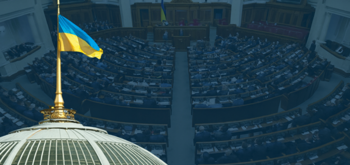Інтер’єр парламентської зали, де сидять численні учасники, ймовірно, народні депутати. Розташування — півколом, з робочими місцями, столами та центральною трибуною. На передньому плані — купол із золотим шпилем, увінчаним синьо-жовтим прапором України. У залі також видно державний прапор. Обстановка — офіційна, урядова, що символізує законодавчу діяльність у Верховній Раді України.