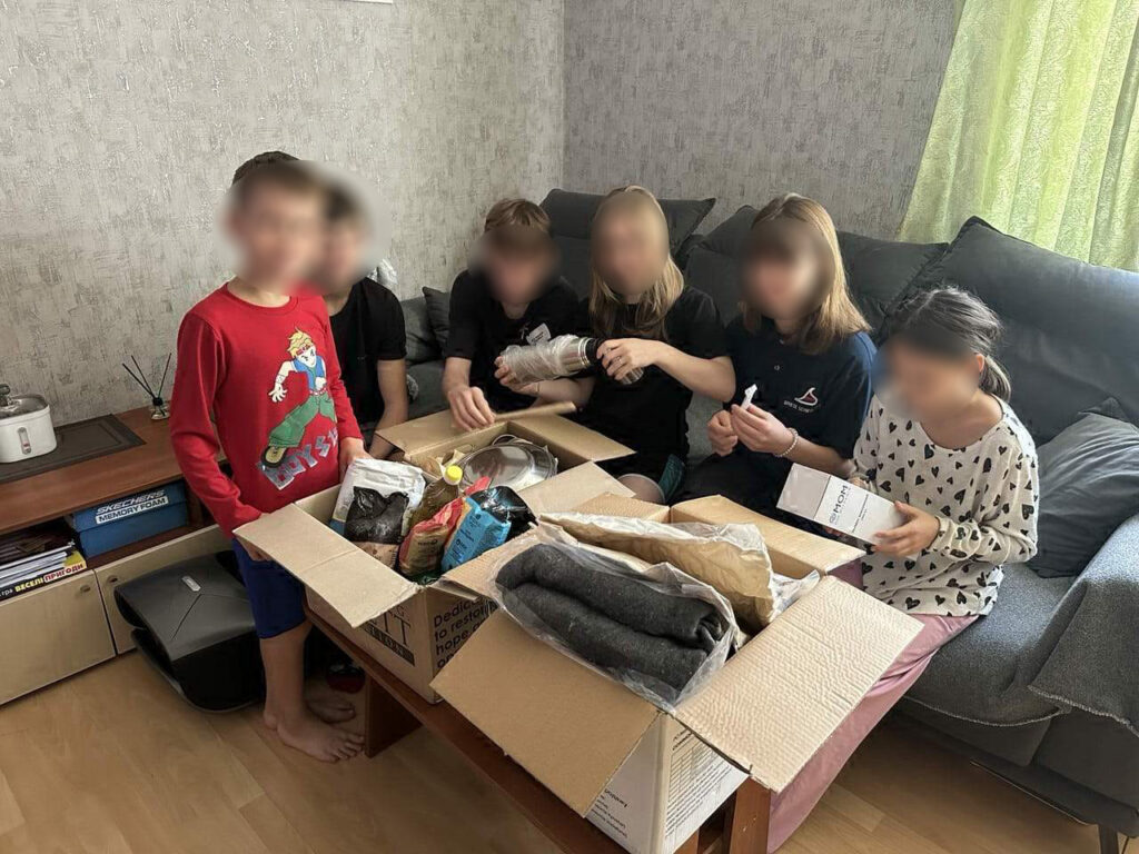 У кімнаті з м’яким освітленням шестеро дітей зібралися навколо столу, розглядаючи вміст двох відкритих картонних коробок. У коробках — продукти, одяг і побутові речі. Один хлопчик стоїть, решта сидять на дивані, уважно досліджуючи речі. На задньому плані — диван з подушками, столик із рисоваркою та ароматичними паличками, а також полиця з коробками, підписаними «MEMORY FOAM» і «RECLINER OTTOMAN».