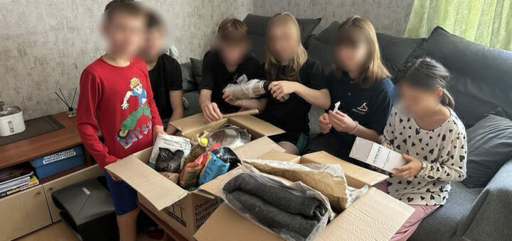 У кімнаті з м’яким освітленням шестеро дітей зібралися навколо столу, розглядаючи вміст двох відкритих картонних коробок. У коробках — продукти, одяг і побутові речі. Один хлопчик стоїть, решта сидять на дивані, уважно досліджуючи речі. На задньому плані — диван з подушками, столик із рисоваркою та ароматичними паличками, а також полиця з коробками, підписаними «MEMORY FOAM» і «RECLINER OTTOMAN».