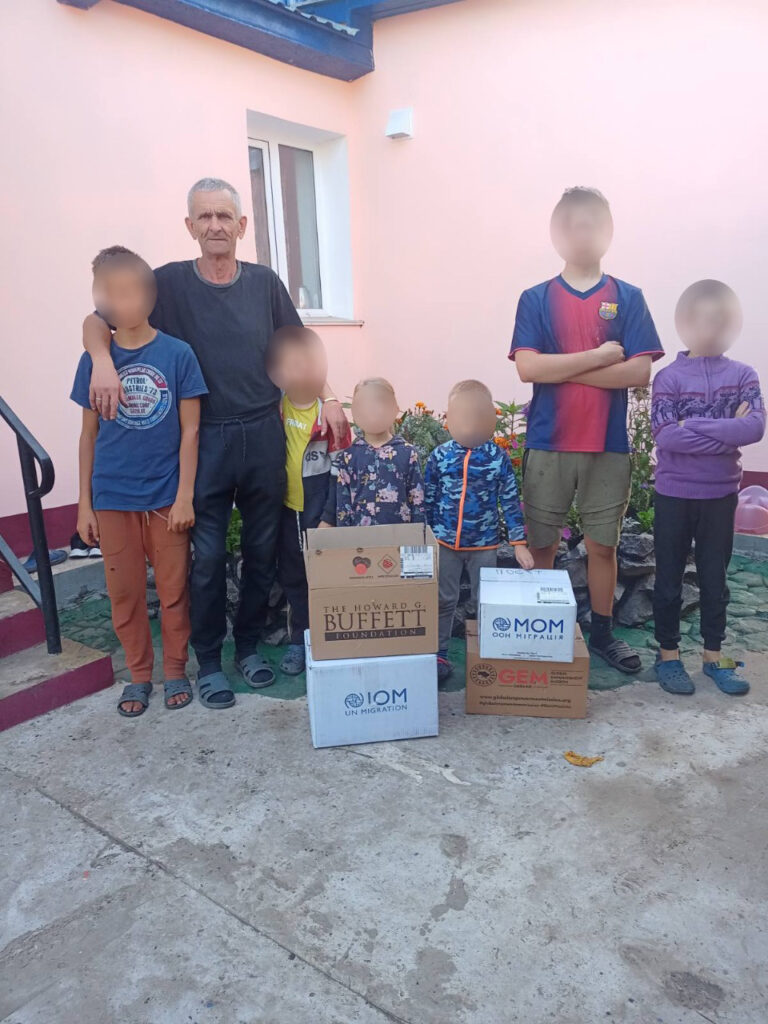 На вулиці перед світло-рожевою будівлею стоїть семеро людей: один старший і шестеро молодших. Їхні обличчя розмиті для конфіденційності. Вони розташовані позаду трьох картонних коробок, що лежать на землі. На коробках видно логотипи міжнародних організацій, зокрема «IOM UN MIGRATION», «THE HOWARD G. BUFFETT FOUNDATION» та «Global Empowerment Mission».