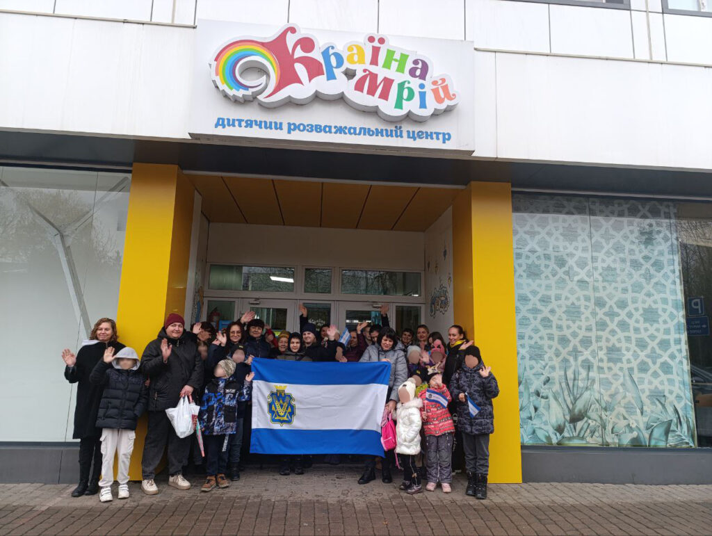 Група людей, серед яких діти та дорослі, стоять перед входом до будівлі з яскравою назвою Країна мрій". Багато учасників усміхаються та махають руками. В центрі групи — прапор синьо-білого кольору з жовто-синім емблемою. 