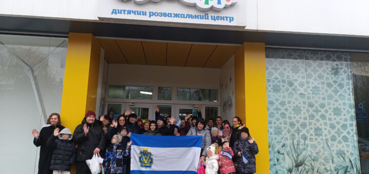 Група людей, серед яких діти та дорослі, стоять перед входом до будівлі з яскравою назвою Країна мрій". Багато учасників усміхаються та махають руками. В центрі групи — прапор синьо-білого кольору з жовто-синім емблемою.