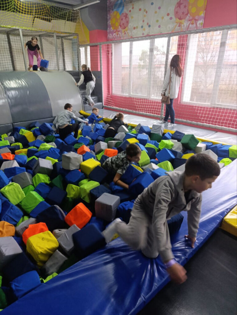 У приміщенні дитячого майданчика кілька дітей активно граються серед м’яких кубів різних кольорів — синіх, зелених, жовтих, оранжевих, сірих і білих. Деякі діти лазять по м’якій конструкції, інші стрибають або повзають між кубами. Простір обгороджений захисною сіткою, а великі вікна пропускають природне світло. Біля вікон стоїть жінка, яка спостерігає за дітьми. Стіни прикрашені яскравими кольорами та святковими візерунками, що створює веселу та безпечну атмосферу.