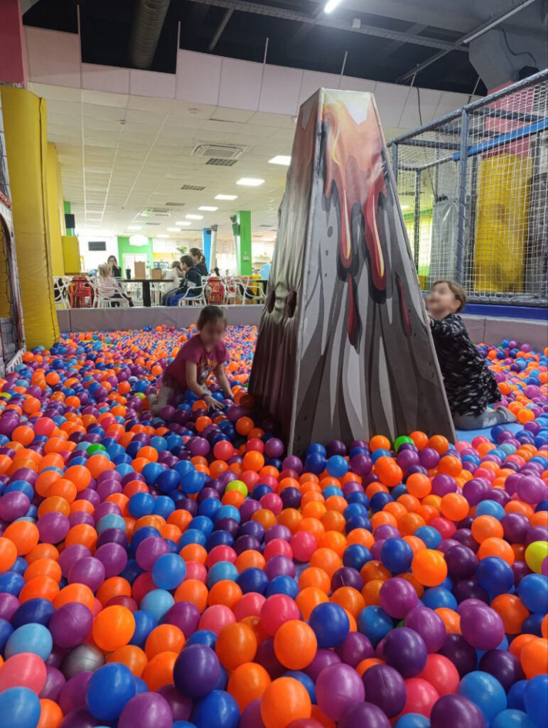 У приміщенні дитячого розважального центру — басейн із кольоровими пластиковими кульками. У центрі — велика конструкція у формі вулкана з принтом, що імітує потоки лави. Двоє дітей граються поруч із вулканом серед кульок. На задньому плані — зона з столами, де сидять дорослі. Інтер’єр яскравий, створений для активних ігор та відпочинку дітей.