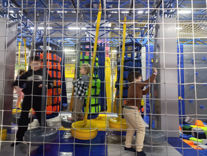 У приміщенні дитячого ігрового центру кілька дітей граються всередині огородженої зони з підвісними м’якими циліндрами. Циліндри мають кольорові сегменти — оранжеві, зелені та сині — і слугують для лазіння або проходження перешкод. Підлога м’яка, для безпеки. Праворуч видно вертикальну стіну з виступами для лазіння. Конструкції зроблені з металу та захисних сіток. Освітлення яскраве, атмосфера активна й стимулююча.