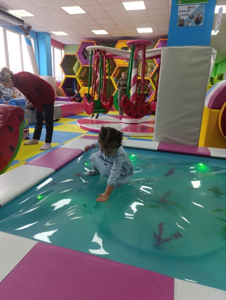 Приміщення дитячого ігрового майданчика з яскравими м’якими конструкціями. На передньому плані — маленька дитина взаємодіє з прозорою поверхнею, під якою видно декоративні водяні рослини, що створює ілюзію ставка. Навколо — рожеві та білі м’які елементи. У глибині — шестикутні конструкції для лазіння, гойдалка з сидіннями у вигляді кавунів, а також кілька дорослих і дітей, які граються.