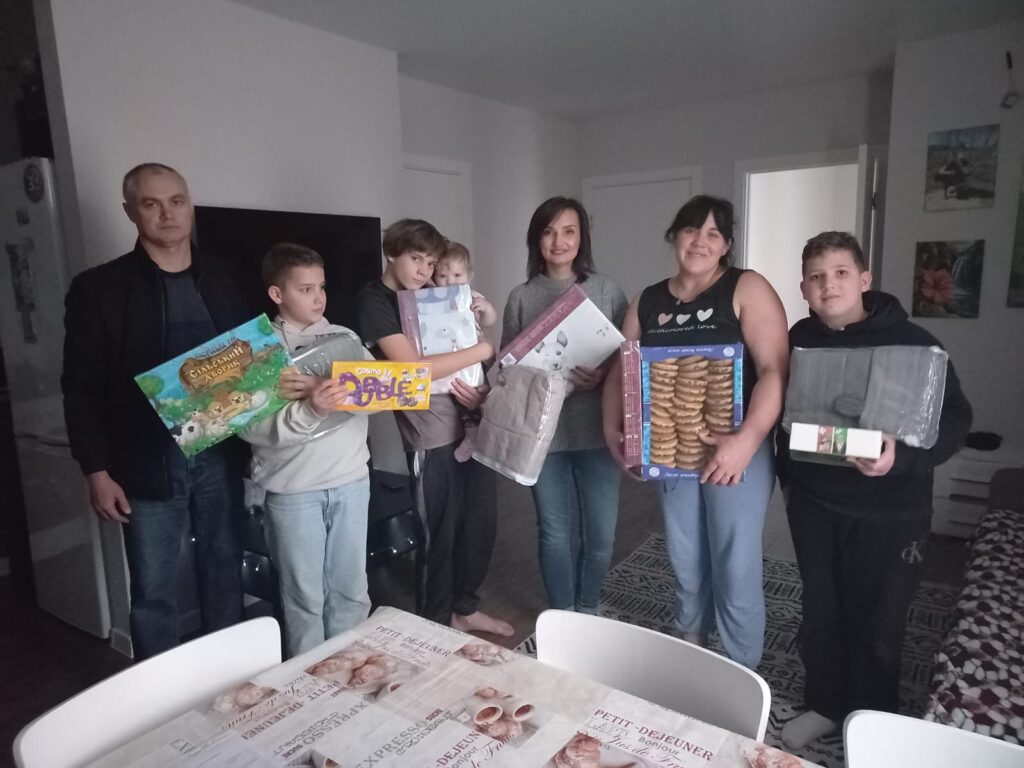 Сім людей різного віку позують для фото в домашньому інтер’єрі. Вони тримають настільні ігри, печиво, ковдру та інші паковані речі. Видно меблі — обідній стіл зі скатертиною з французькими написами «PETIT DEJEUNER» і «EXPRESSO», стільці, картини на стінах.