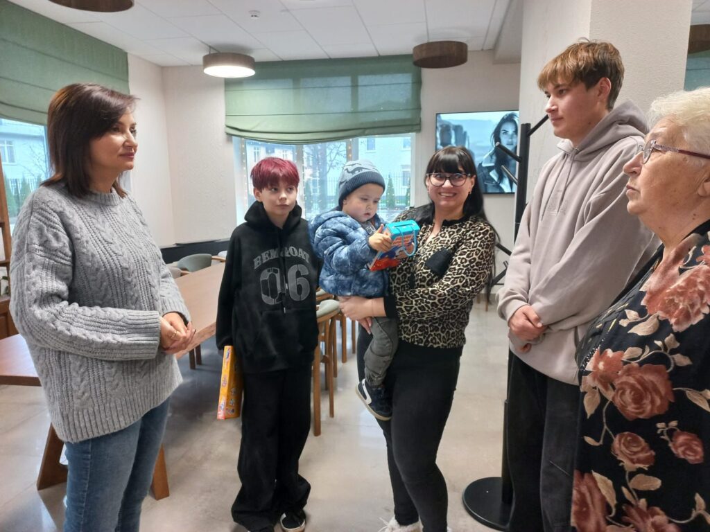 У світлій кімнаті з великими вікнами та зеленими жалюзі стоїть шестеро людей. Серед них — жінка в сірому светрі, дитина з рудим волоссям, яка тримає коробку, жінка в леопардовій блузі з малюком у синьому одязі, юнак у світлій худі та літня жінка у квітчастій блузі. Позаду — столи й стільці, на стіні — чорно-білий портрет.