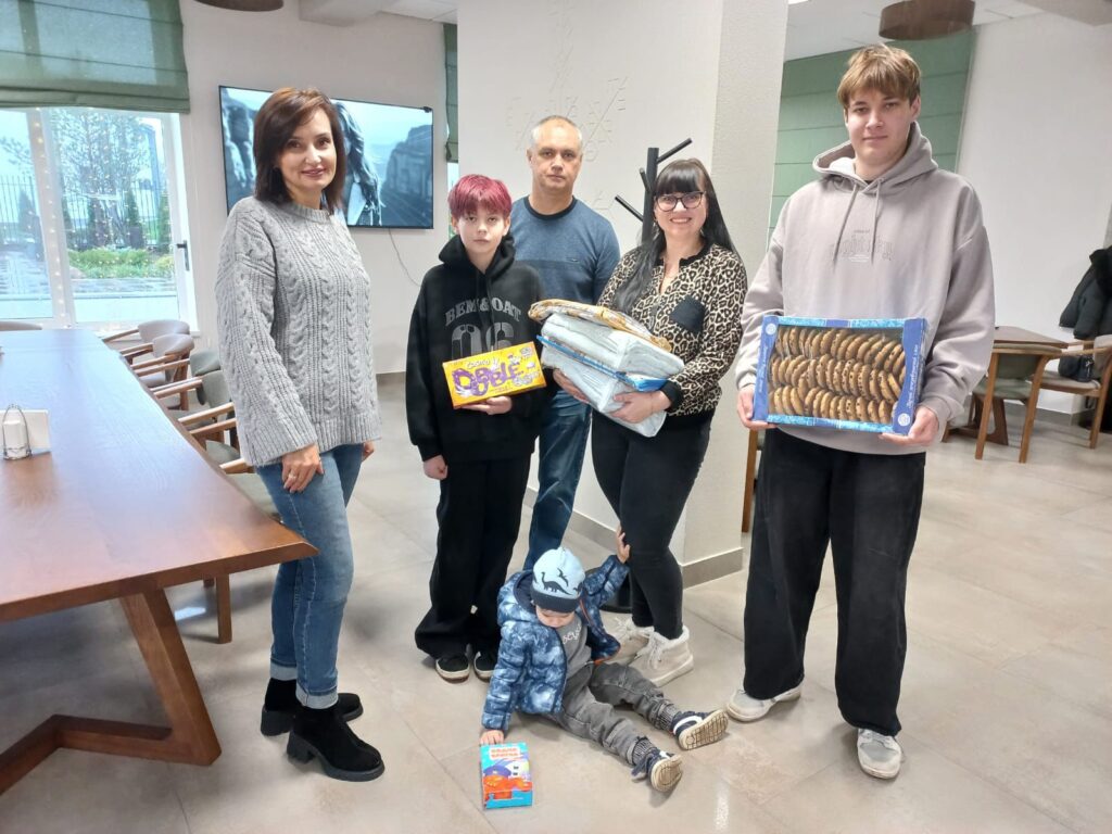 У сучасному приміщенні стоїть група з шести людей — четверо дорослих і двоє дітей. Один дорослий тримає складені ковдри, одна дитина — коробку печива «Domaćica», інша — коробку снеків «Bobi». Маленька дитина сидить на підлозі й грається з коробкою. Позаду — столи, стільці та телевізор на стіні, який показує чорно-біле зображення