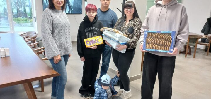 У сучасному приміщенні стоїть група з шести людей — четверо дорослих і двоє дітей. Один дорослий тримає складені ковдри, одна дитина — коробку печива «Domaćica», інша — коробку снеків «Bobi». Маленька дитина сидить на підлозі й грається з коробкою. Позаду — столи, стільці та телевізор на стіні, який показує чорно-біле зображення