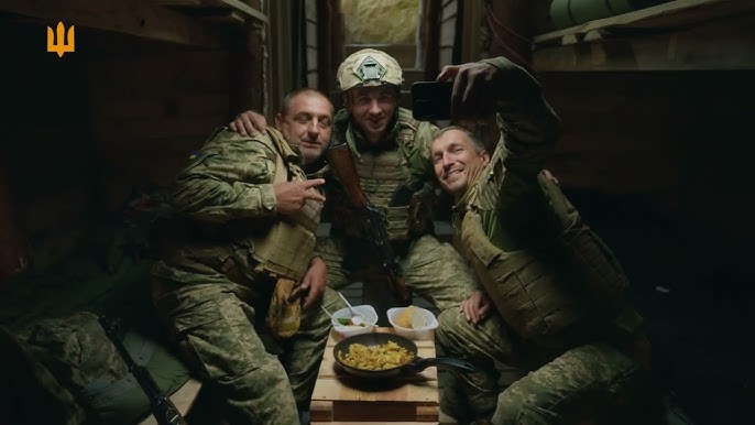 Троє військових у камуфляжній формі сидять разом у дерев’яній споруді, схожій на бліндаж. Один із них тримає смартфон і робить селфі. Всі усміхнені. Перед ними — стіл із сковорідкою та мисками, що свідчить про спільну трапезу. Позаду — двоярусні ліжка та військове спорядження. У верхньому лівому куті — жовтий тризуб.