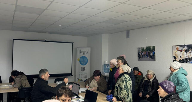 У великій кімнаті зібралося кілька людей. Частина сидить за довгим столом із ноутбуками, блокнотами та синіми прапорцями, інші — уздовж стіни, можливо, очікують допомоги або беруть участь у розмові. У приміщенні — стеля з вбудованим освітленням, екран для проєктора та плакати або фотографії на стінах. На задньому плані — банер із написом «ШоТрик», круглою емблемою та QR-кодом.