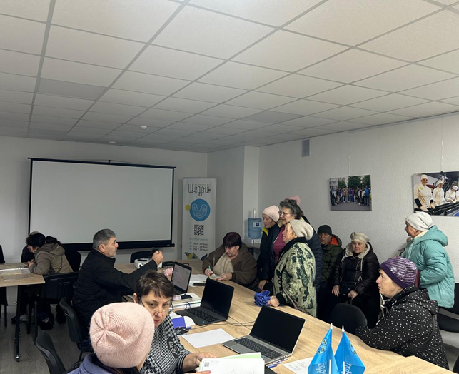 У великій кімнаті зібралося кілька людей. Частина сидить за довгим столом із ноутбуками, блокнотами та синіми прапорцями, інші — уздовж стіни, можливо, очікують допомоги або беруть участь у розмові. У приміщенні — стеля з вбудованим освітленням, екран для проєктора та плакати або фотографії на стінах. На задньому плані — банер із написом «ШоТрик», круглою емблемою та QR-кодом. 