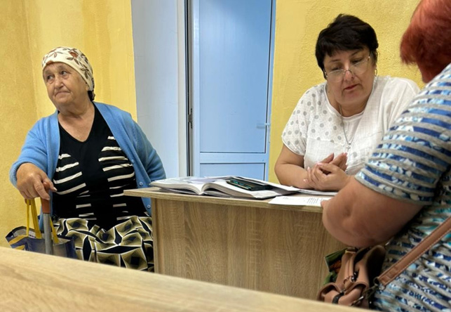 У кімнаті з жовтими стінами троє жінок беруть участь у розмові. Ліворуч — жінка в хустці, синьому кардигані та сукні з геометричним орнаментом, тримає тростину й уважно слухає. У центрі — жінка за дерев’яним столом, у білій блузі з крапками, дивиться на відкриті документи. Праворуч — жінка з рудим волоссям у синьо-білій смугастій кофті, сидить із сумкою на колінах і звернена до столу. 