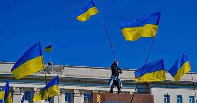 На фотографії зображено святкову сцену на тлі урядової будівлі. У центрі — людина стоїть на підвищенні, тримаючи в руках великий прапор України. Навколо — багато українських прапорів, які майорять у повітрі. Небо ясне, синє, що підкреслює яскраві кольори прапорів.