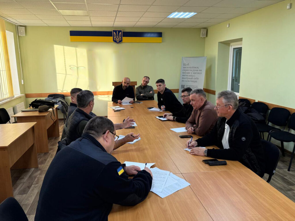 На фото — десятеро людей за великим столом у світлій кімнаті з українським прапором і гербом на стіні. Усі зосереджені: хтось пише, хтось слухає. На задньому плані — банер із текстом про європейську інтеграцію, що підкреслює офіційний характер зустрічі