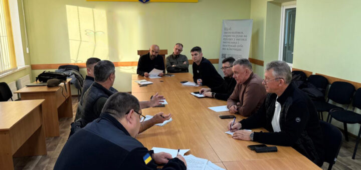 На фото — десятеро людей за великим столом у світлій кімнаті з українським прапором і гербом на стіні. Усі зосереджені: хтось пише, хтось слухає. На задньому плані — банер із текстом про європейську інтеграцію, що підкреслює офіційний характер зустрічі