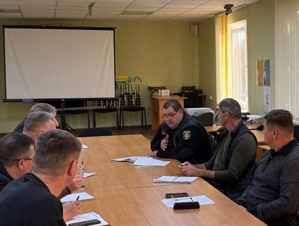Сім чоловіків сидять за столом у світлій кімнаті з проєктором. Один із них — у формі з шевроном — говорить, решта слухають або роблять нотатки. На столі — документи, телефони, ручки.