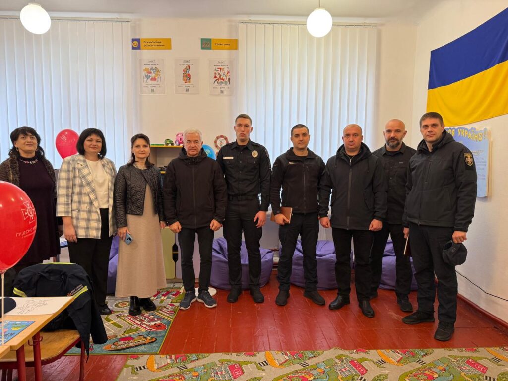 У кімнаті з яскравими дитячими декораціями стоять восьмеро людей, п’ятеро з яких — у чорній формі, ймовірно, представники правоохоронних органів. Решта — у цивільному одязі. На стінах — дитячі малюнки, прапор України, а на підлозі — кольоровий килим