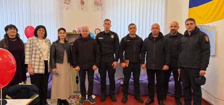 У кімнаті з яскравими дитячими декораціями стоять восьмеро людей, п’ятеро з яких — у чорній формі, ймовірно, представники правоохоронних органів. Решта — у цивільному одязі. На стінах — дитячі малюнки, прапор України, а на підлозі — кольоровий килим