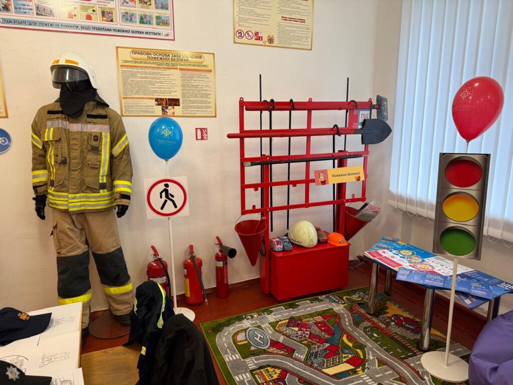 У кімнаті для навчання з безпеки — манекен у формі пожежника, стенд із інструментами (лопати, сокира, каски), кілька вогнегасників, дорожні знаки та макет світлофора. На столі — навчальні матеріали, кепки, повітряні кульки з символікою безпеки. Килим із зображенням доріг і будівель підказує, що простір орієнтований на дітей.