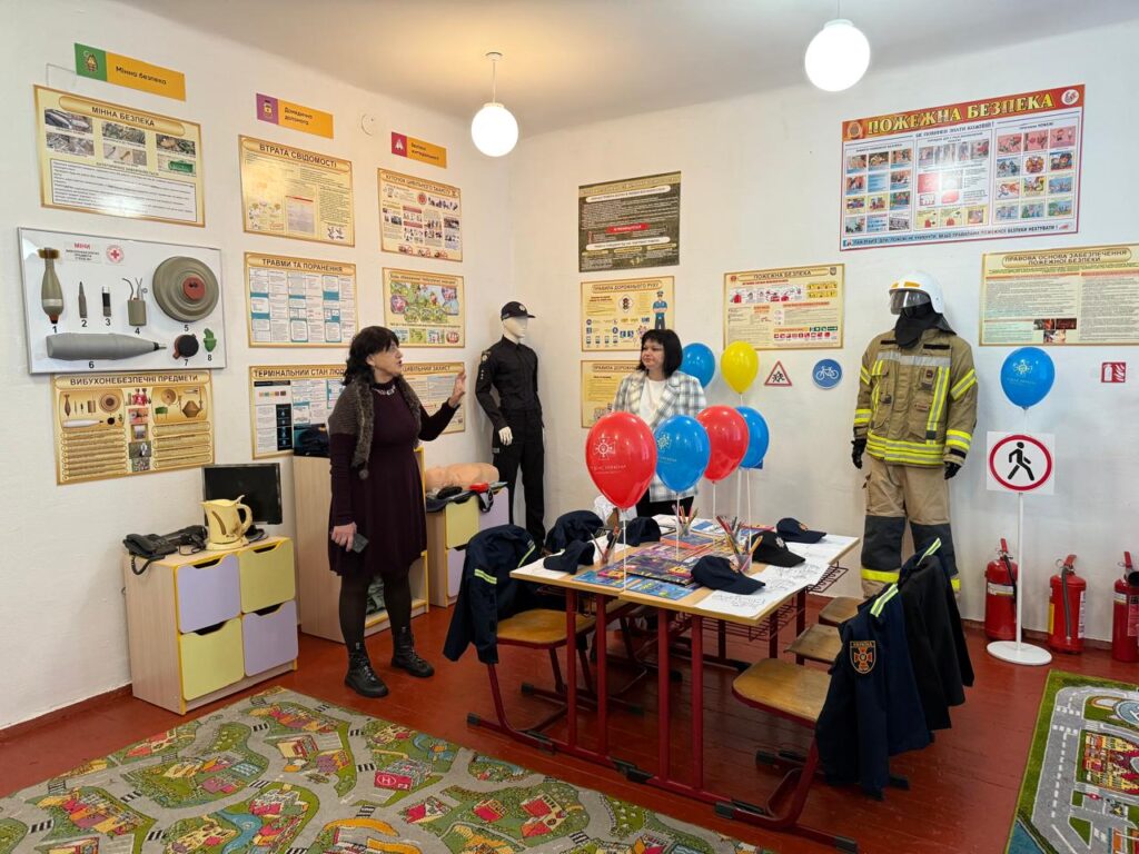 Кімната оформлена як навчальний простір із акцентом на безпеку: на стінах — плакати з пожежної тематики, знаки та схеми евакуації. На столі — повітряні кульки, навчальні матеріали, а поруч — манекени в захисному спорядженні. Килим із зображенням доріг і міста підказує, що заняття орієнтоване на дітей.